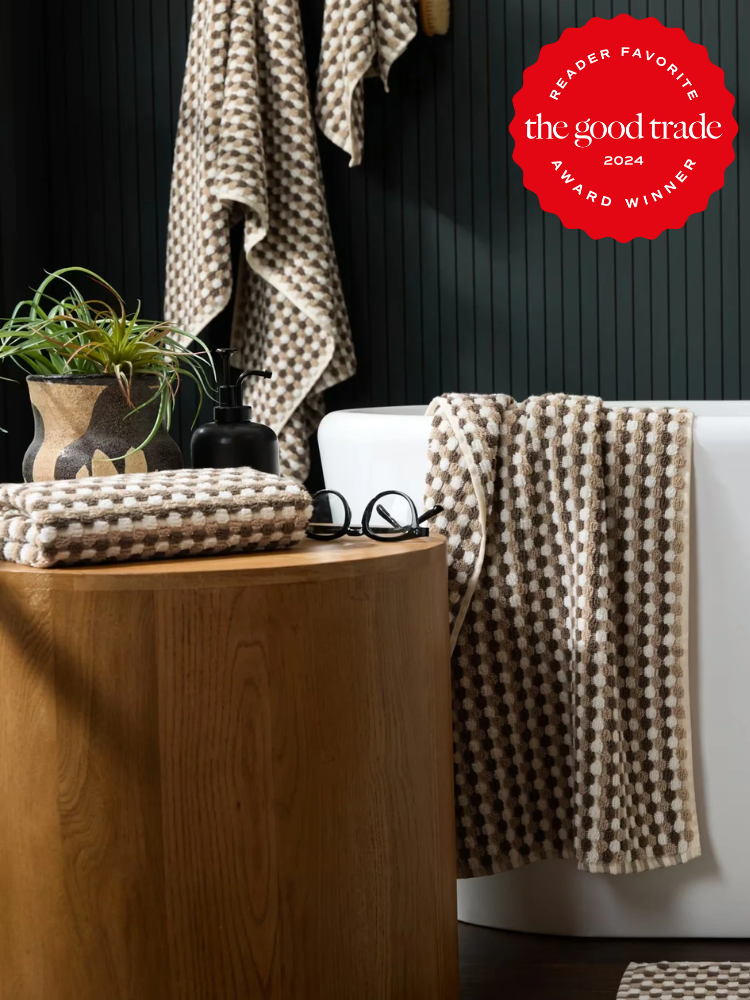 A bathroom with a white tub, wooden side table, stacked and hanging organic checkered towels, potted plant, and a red "The Good Trade 2024 Award Winner" badge in the top right corner.
