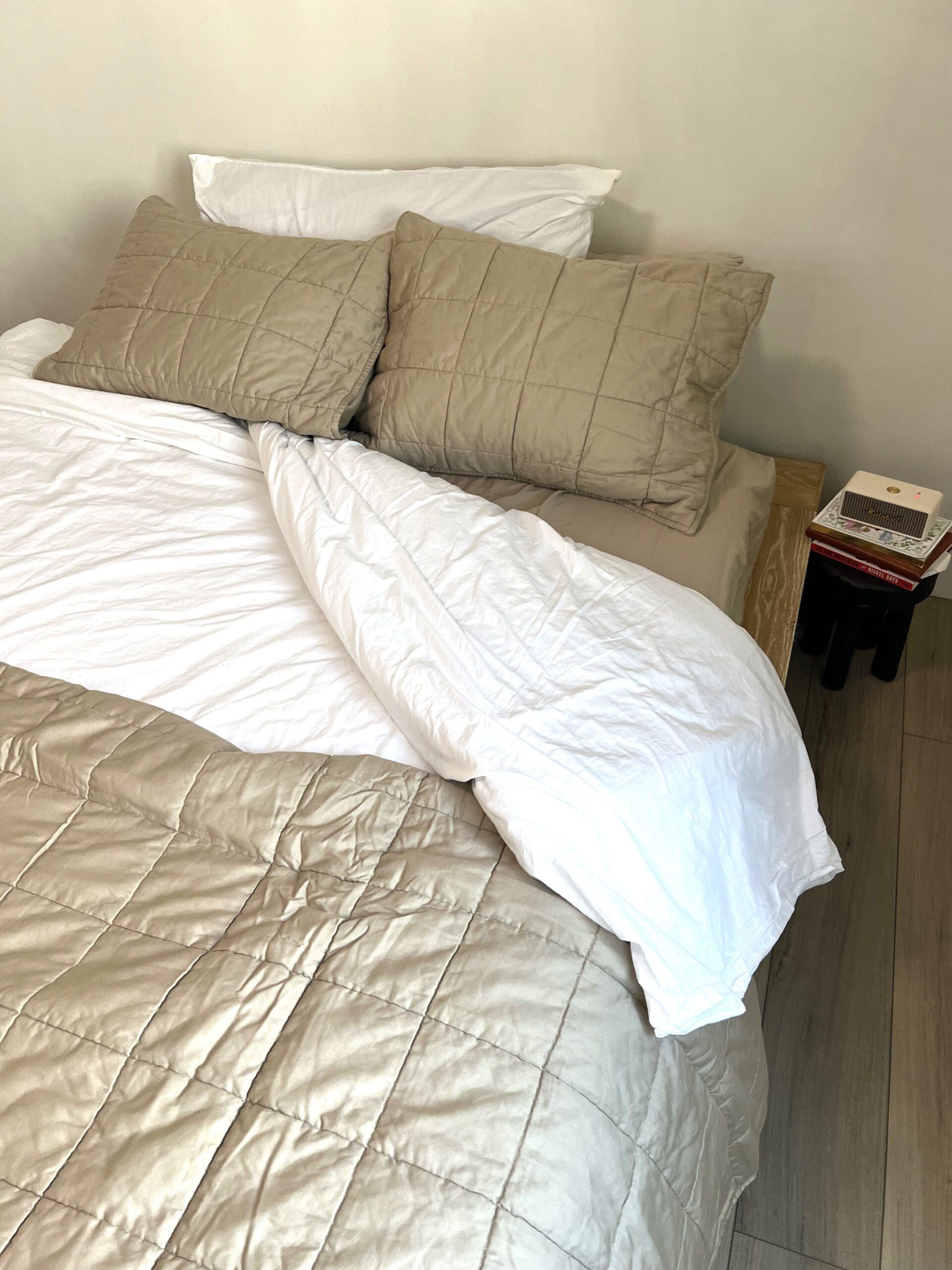 A neatly made bed with beige and white bedding. Two pillows rest against the headboard, and a small table beside the bed holds a stack of books.