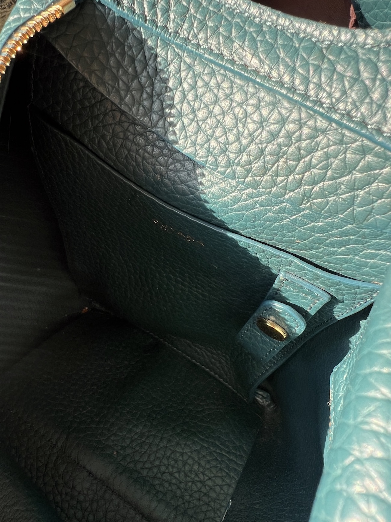 Close-up view of the interior of a teal leather handbag with textured lining, an inner pocket, and a small gold button detail—perfect for anyone searching for an honest Cuyana review.