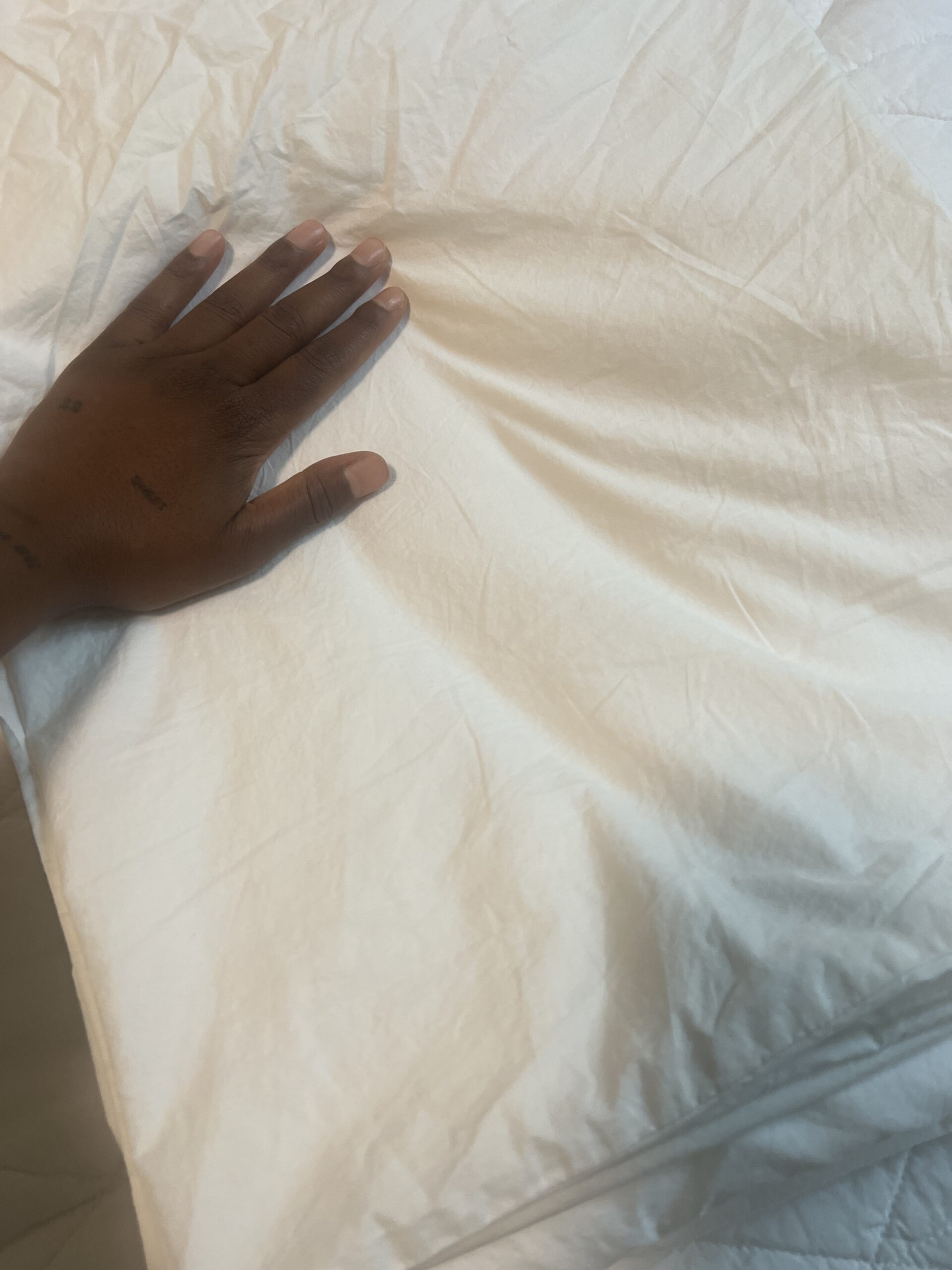 A hand rests on a white, crumpled bedsheet, showcasing skin and fingernails against the textured fabric.