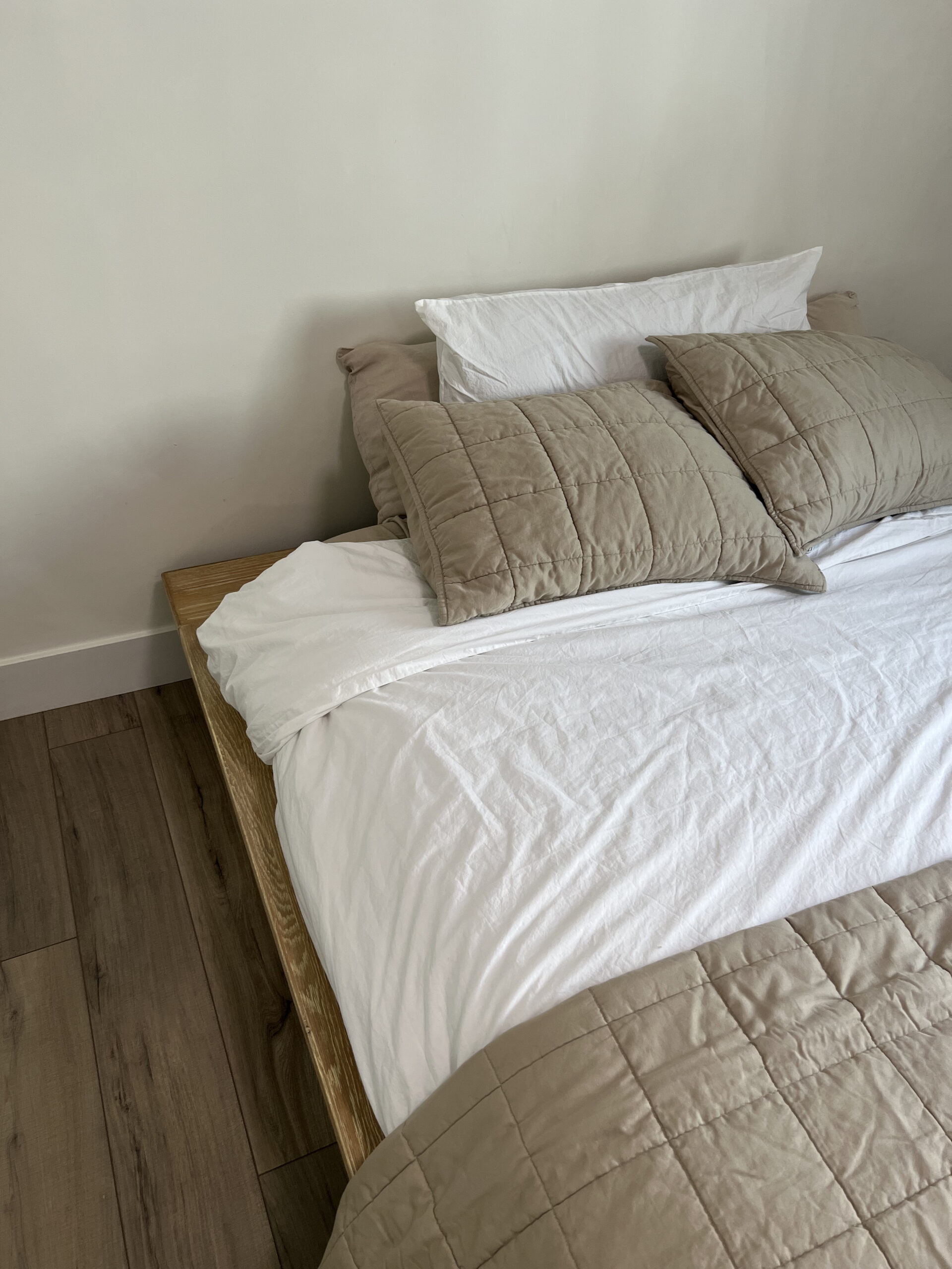 A neatly made bed with a wooden frame features white and beige bedding. The bed is against a white wall and a wooden floor is visible.