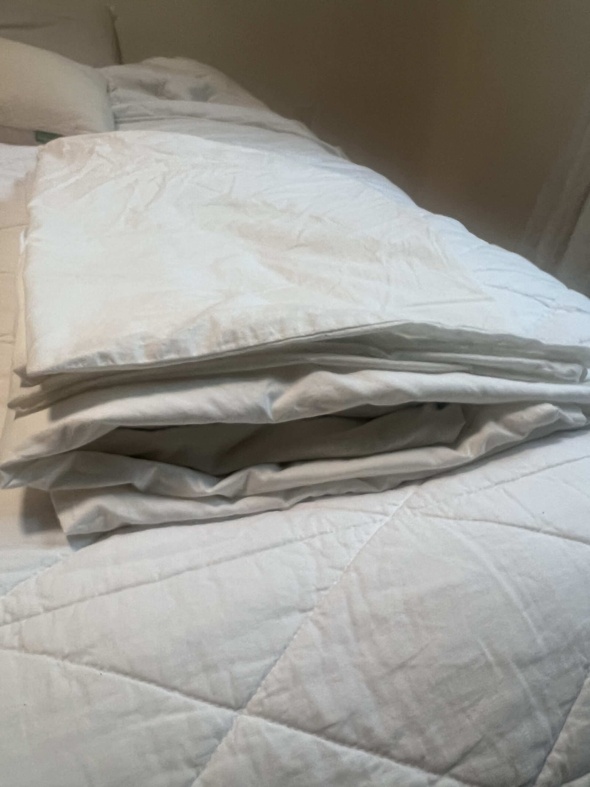 A neatly folded white blanket lays on a white quilted bedspread in a dimly lit room.
