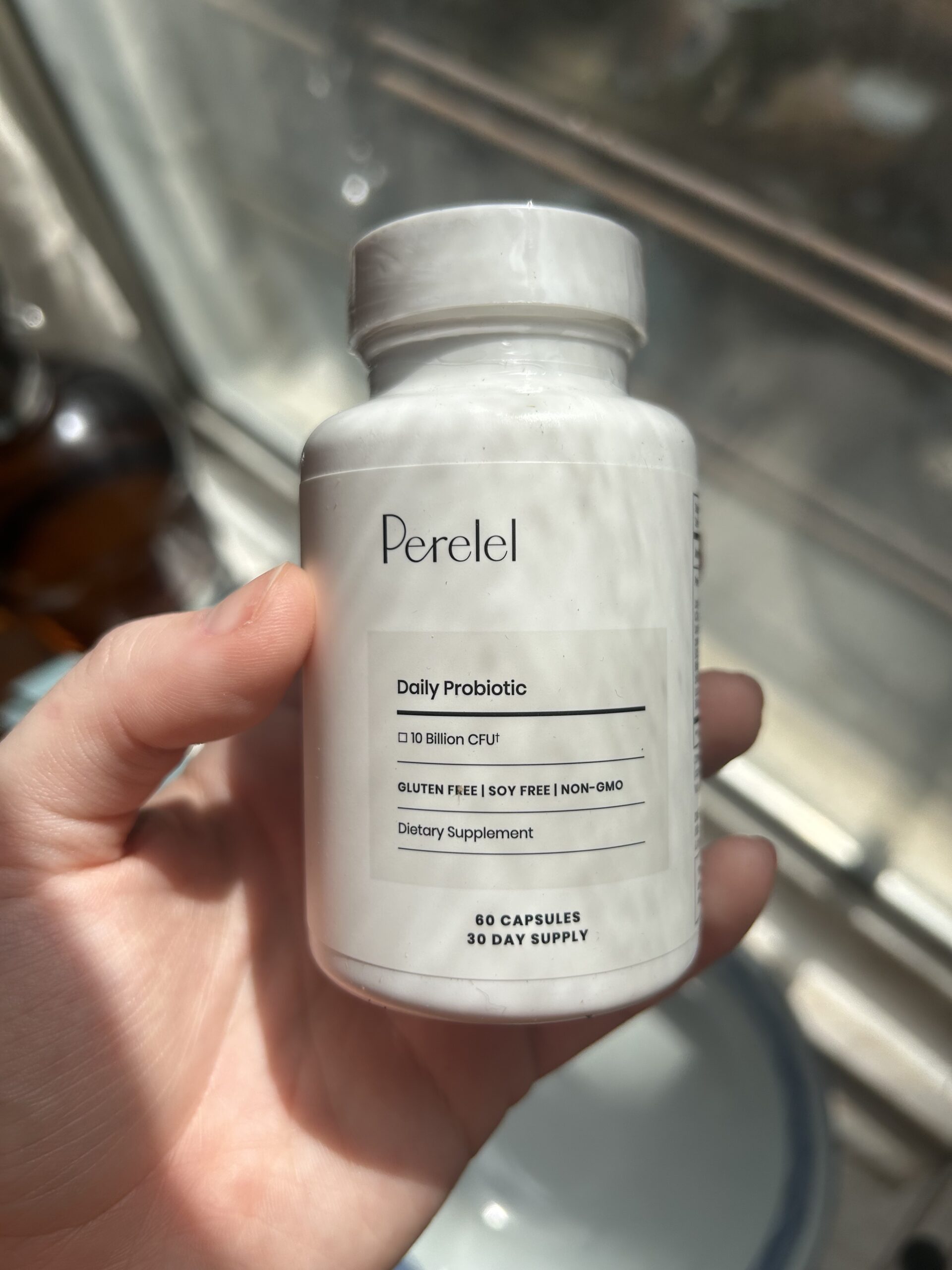 A hand holding a white bottle of Perelel Daily Probiotic dietary supplement, labeled as gluten free, soy free, non-GMO, with 60 capsules for a 30-day supply.