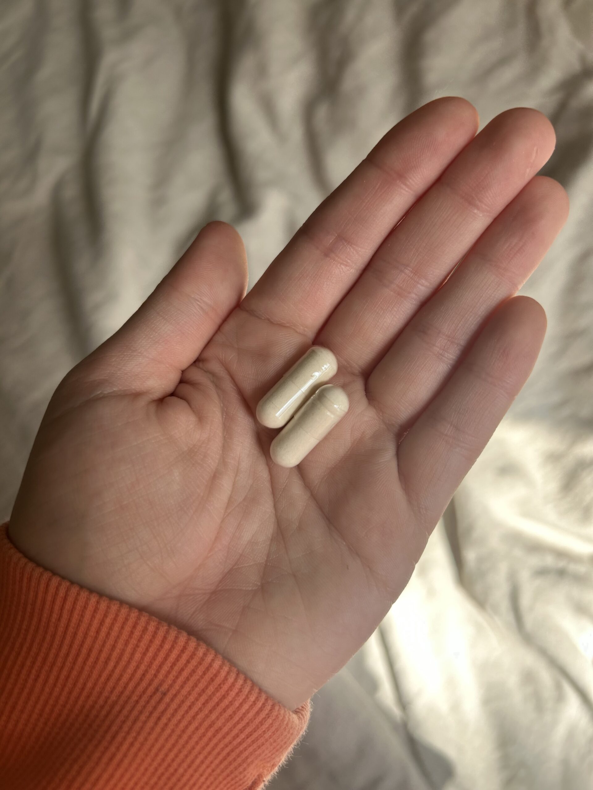 A hand with a peach-colored sleeve holds two white capsules against a light, blurred background.