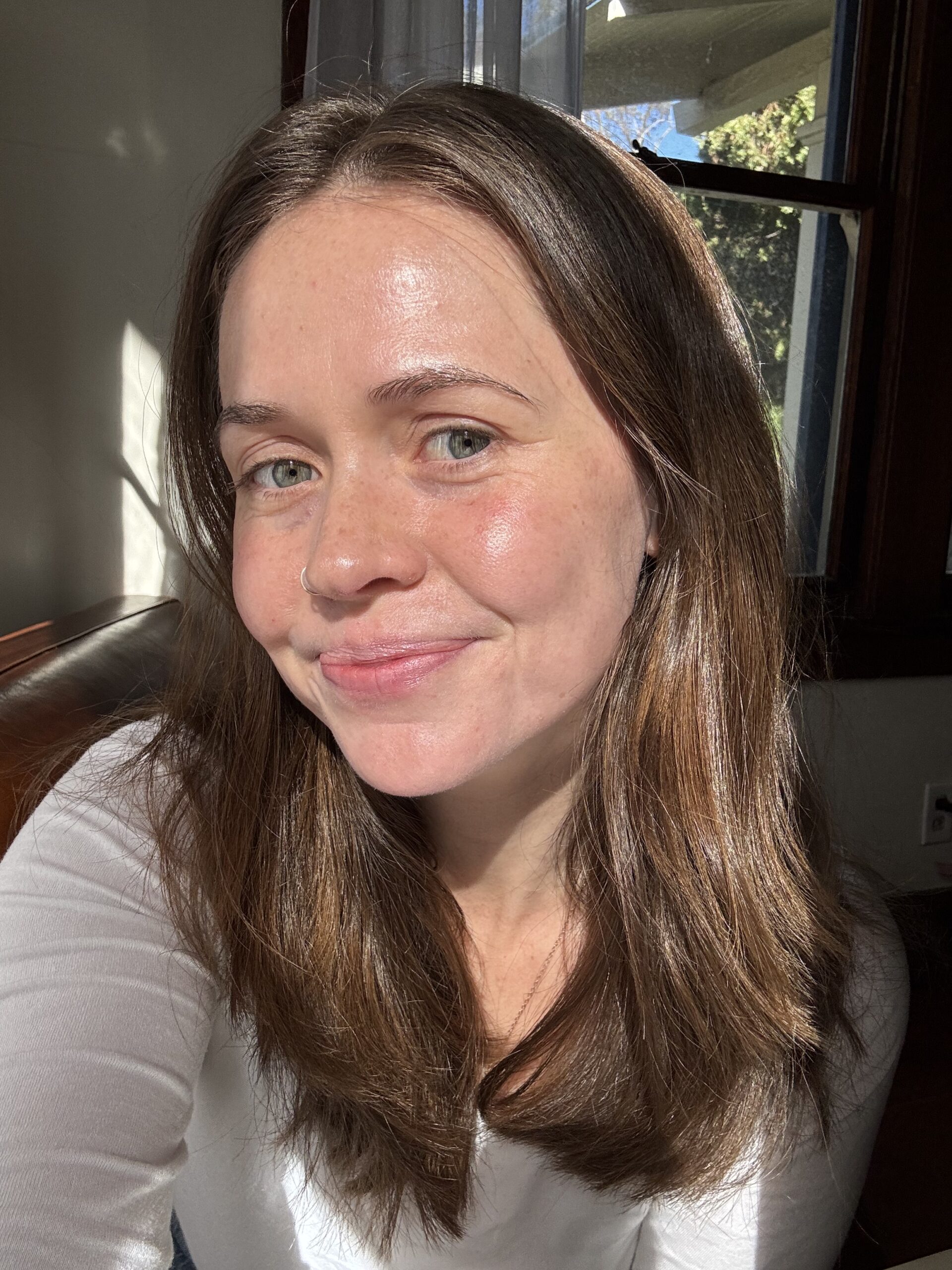 A person with long brown hair and a white shirt smiles at the camera in a sunlit room with a window.