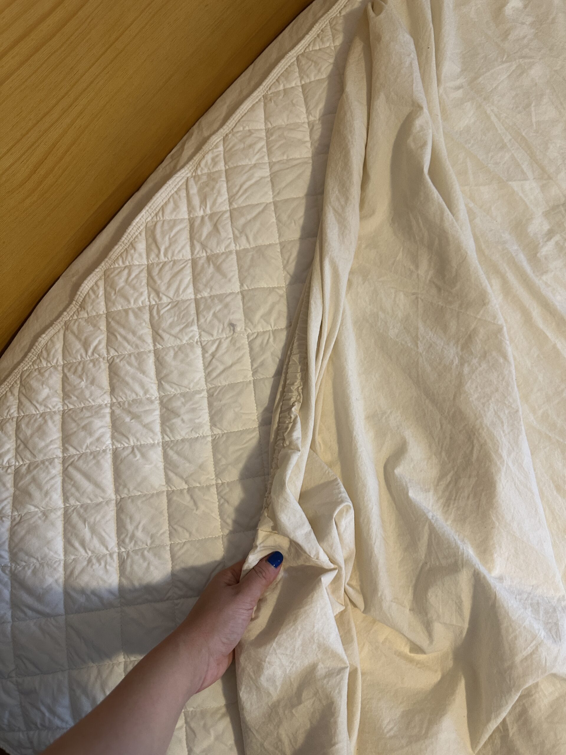 A hand holds the edge of a beige quilt on a wooden surface, revealing its quilted pattern and textured fabric.