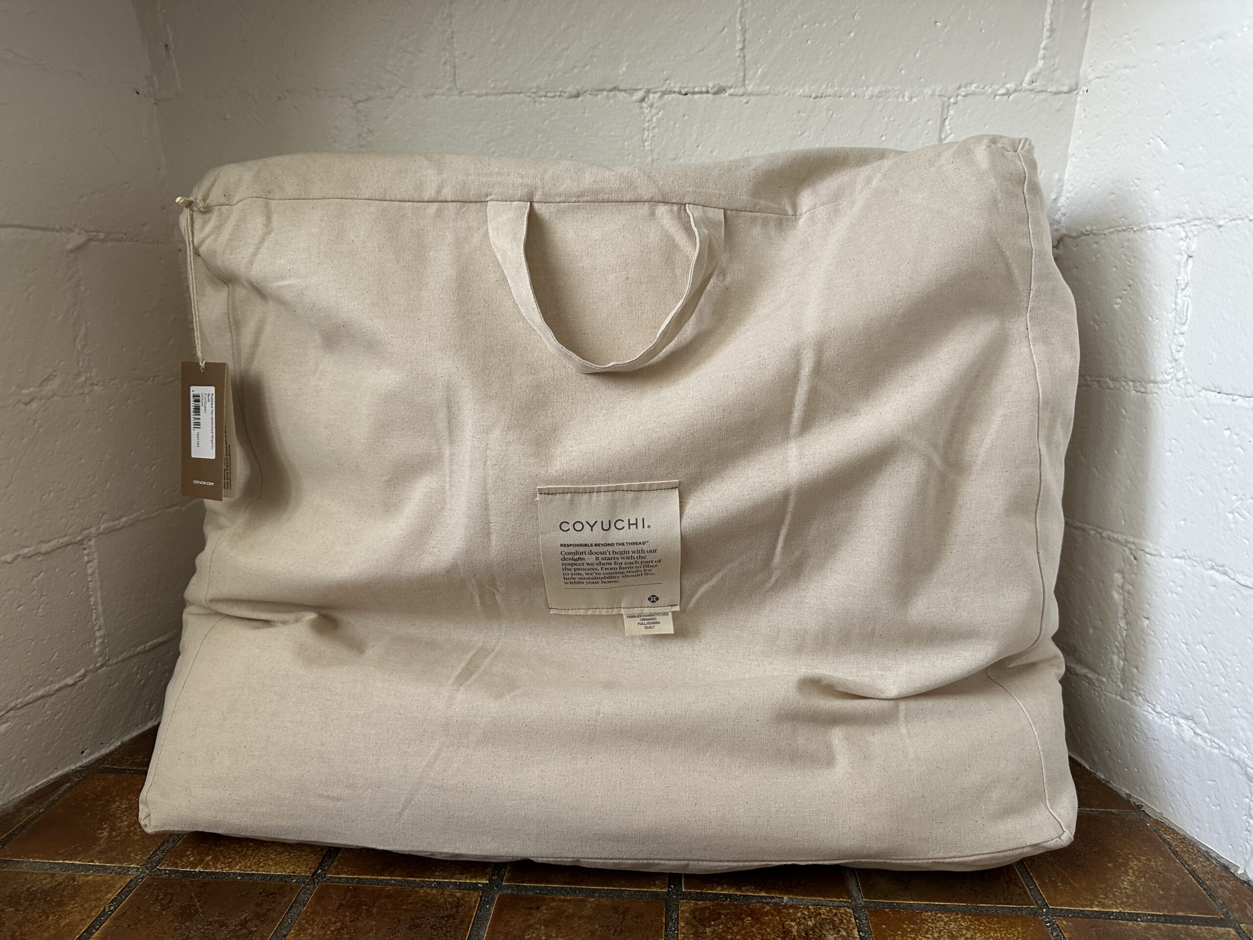 Beige fabric storage bag with handles on a tiled floor. It has a visible tag and brand label on the front.