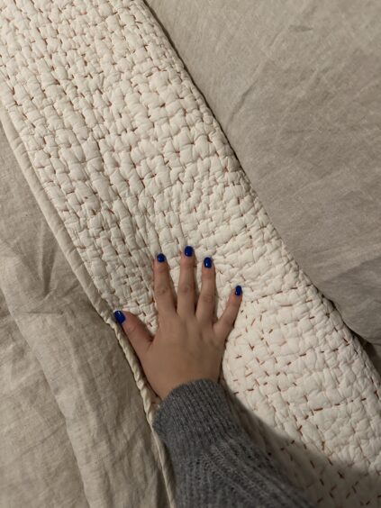 A hand with blue nail polish rests on a beige quilted fabric, surrounded by a textured grey blanket.
