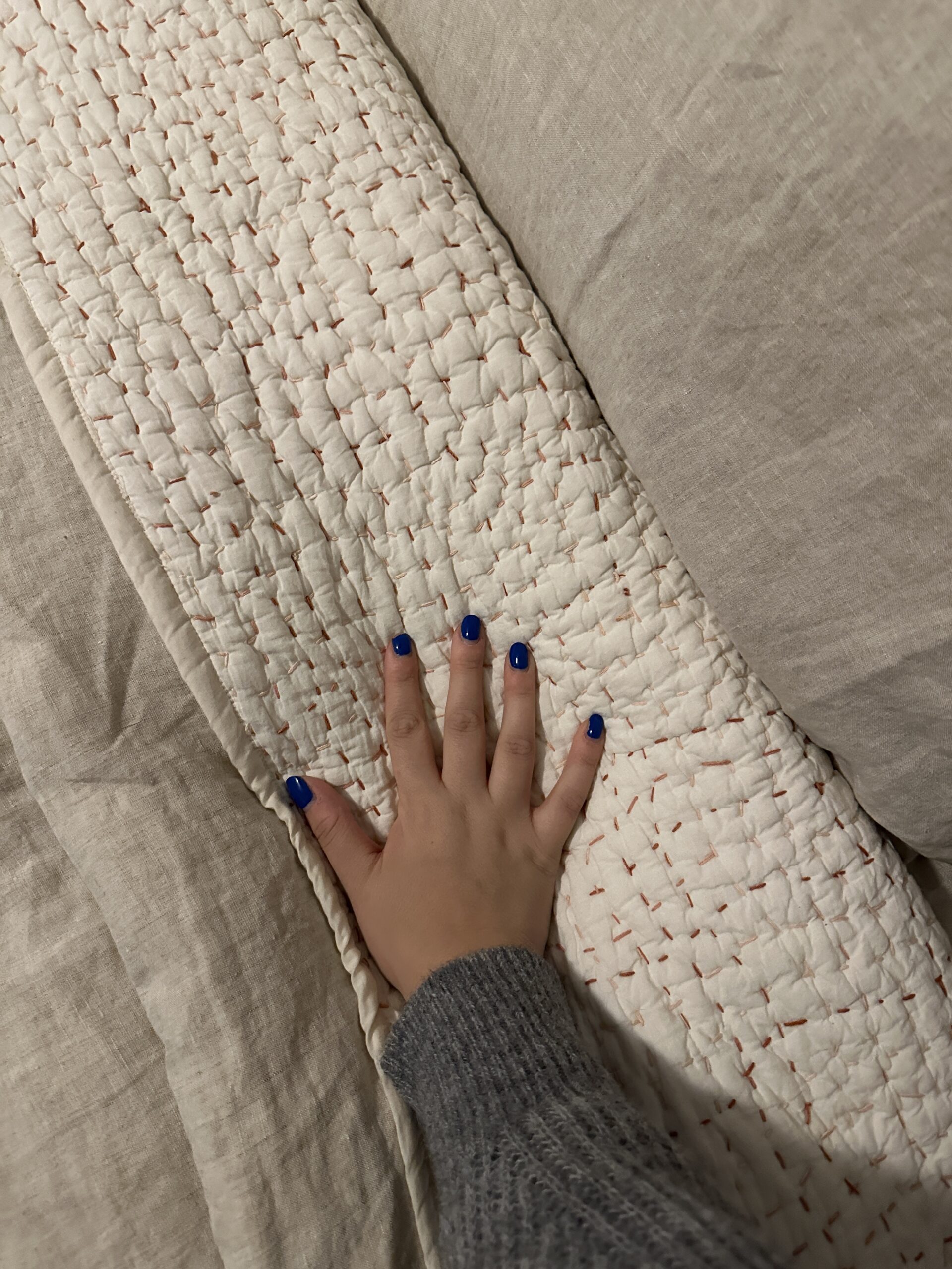 A hand with blue nail polish rests on a beige quilted fabric, surrounded by a textured grey blanket.