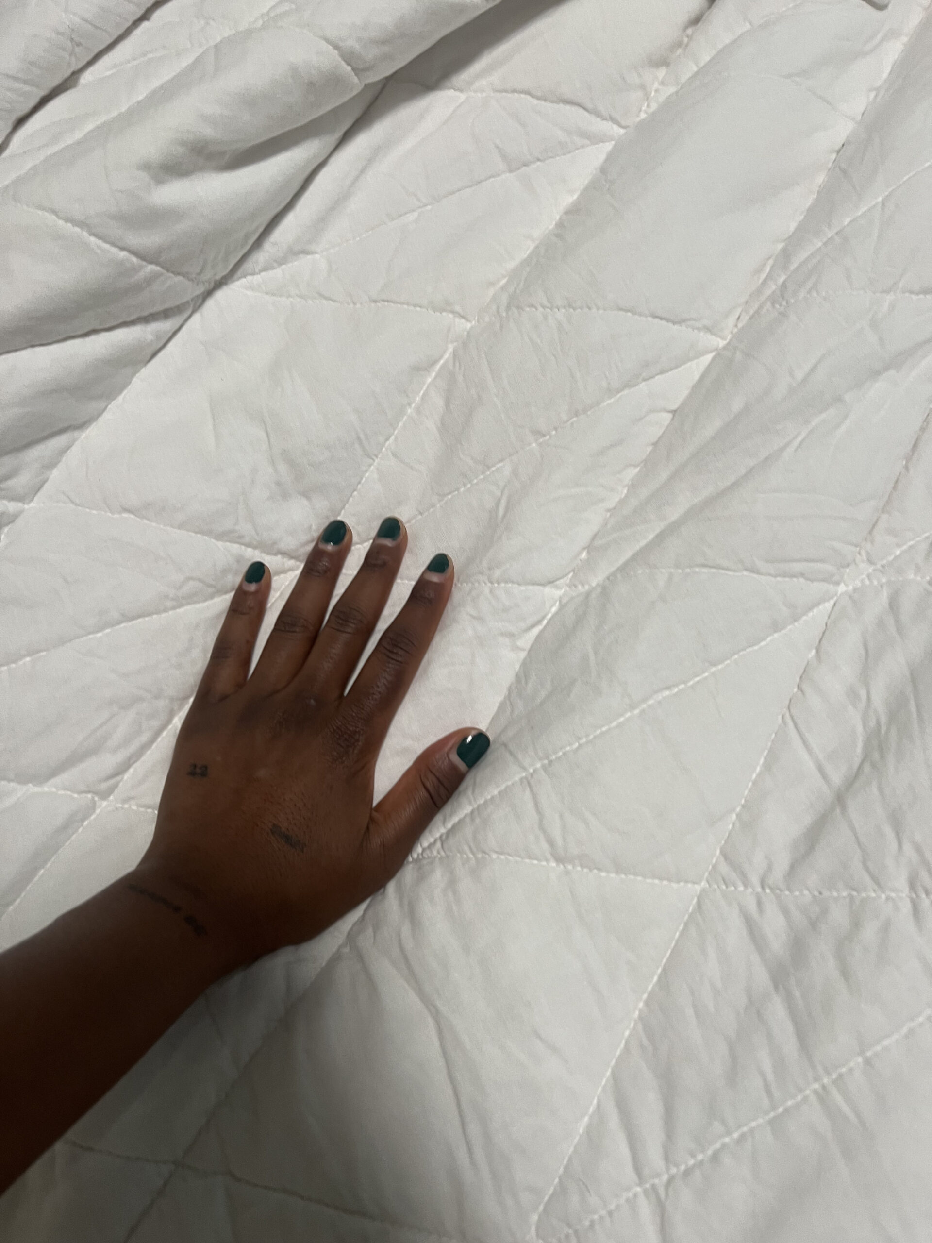 A hand with dark skin and teal-painted nails rests on a white quilted fabric surface.