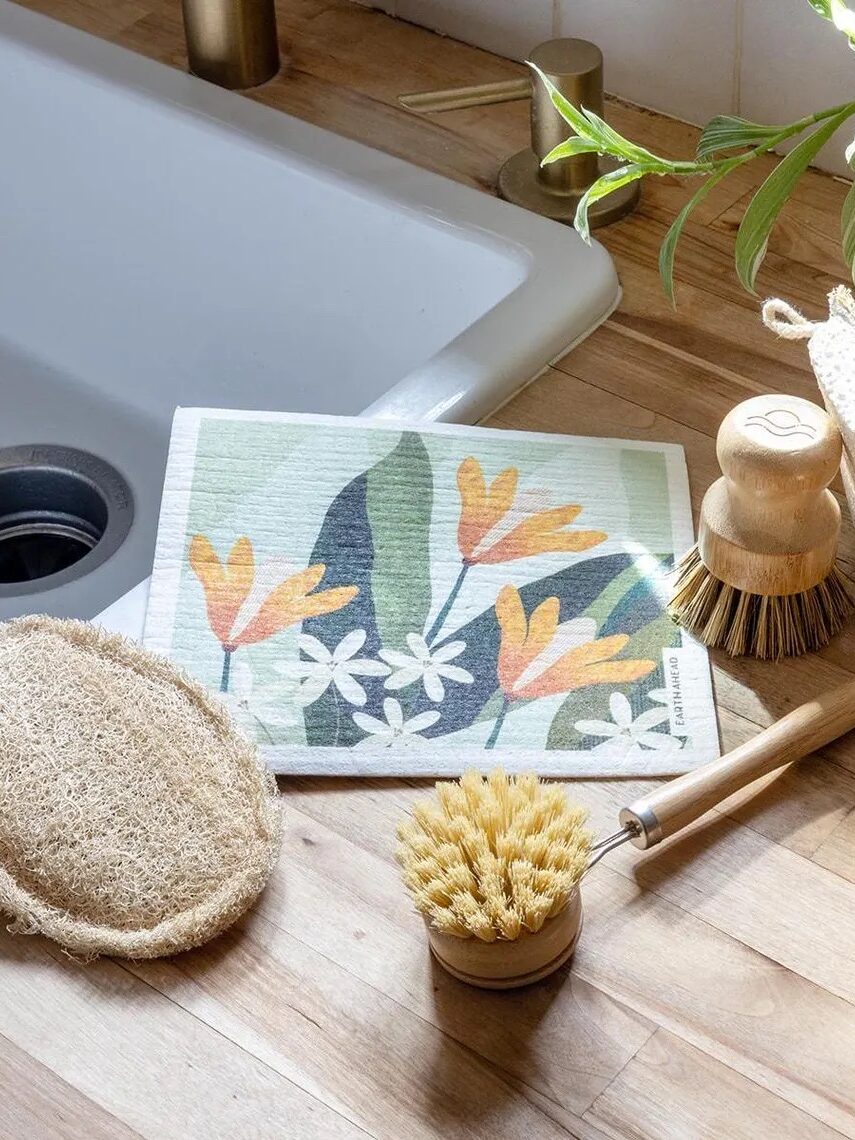 Eco-friendly cleaning tools on a wooden countertop, including brushes and sponges, beside a sink. A small plant is visible in the background.