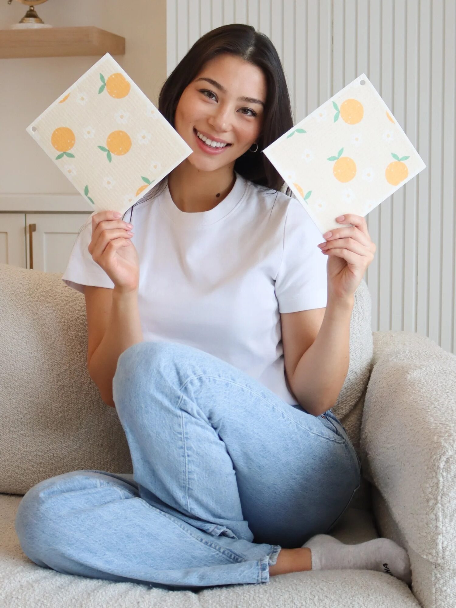 Person sitting on a couch, smiling, holding up two patterned cloths with orange designs. Wearing a white shirt and blue jeans.
