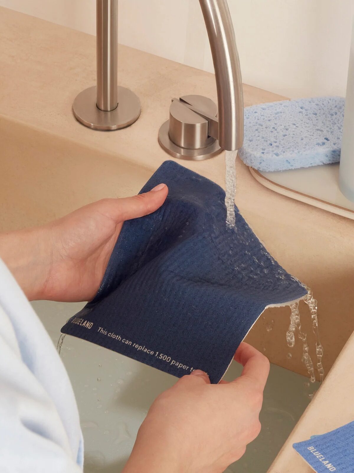 Person holding a blue reusable cloth under running water from a faucet near a bath with light-colored tiles.
