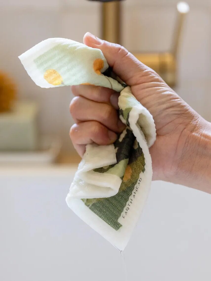 A hand wringing out a colorful cloth over a sink, with water dripping from it.