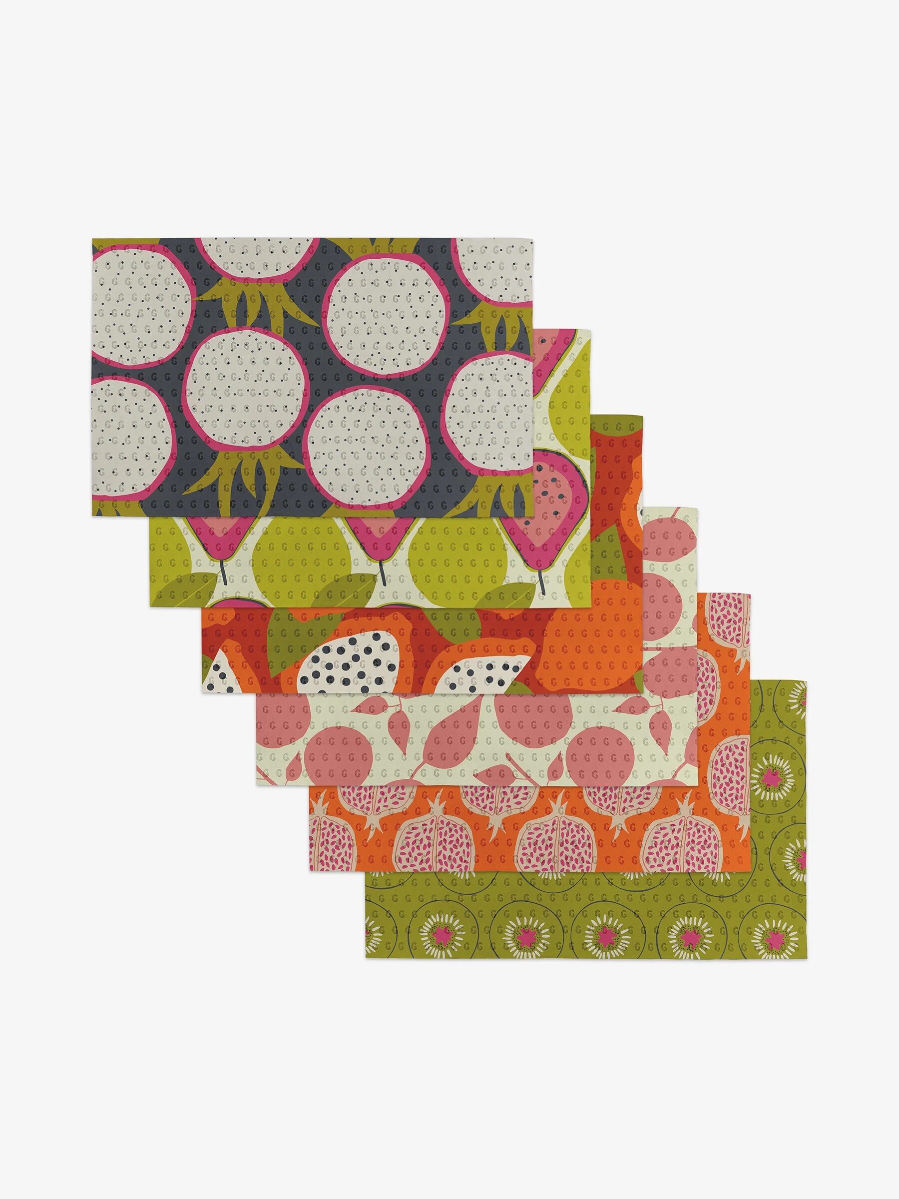 Six patterned placemats are laid out, each with colorful fruit-themed designs, including pomegranates and kiwis, on varied backgrounds.