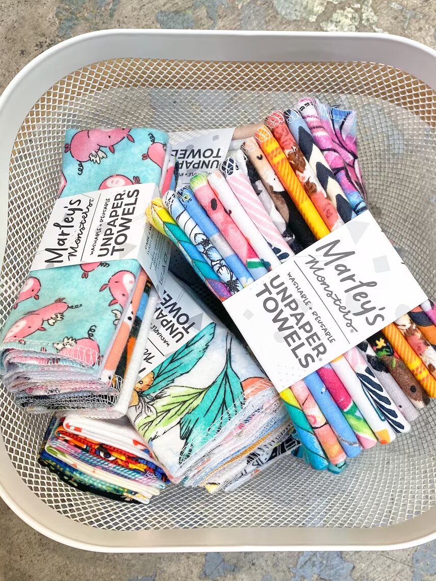A mesh metal basket containing various Marley’s Unpaper Towels with colorful patterns, such as animals and leaves, stacked and packaged with labels.