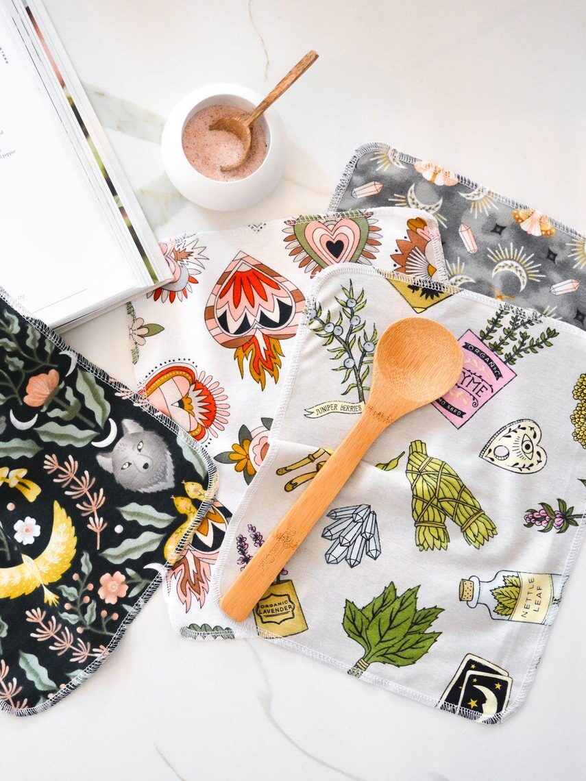 Colorful, patterned fabrics on a surface with a wooden spoon and a bowl of salt, alongside an open book.