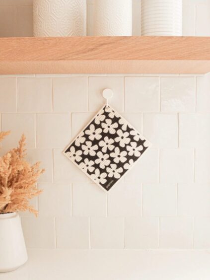 A floral-patterned pot holder hangs on a white tiled kitchen wall with wooden shelves above. A vase with dried plants is on the counter.