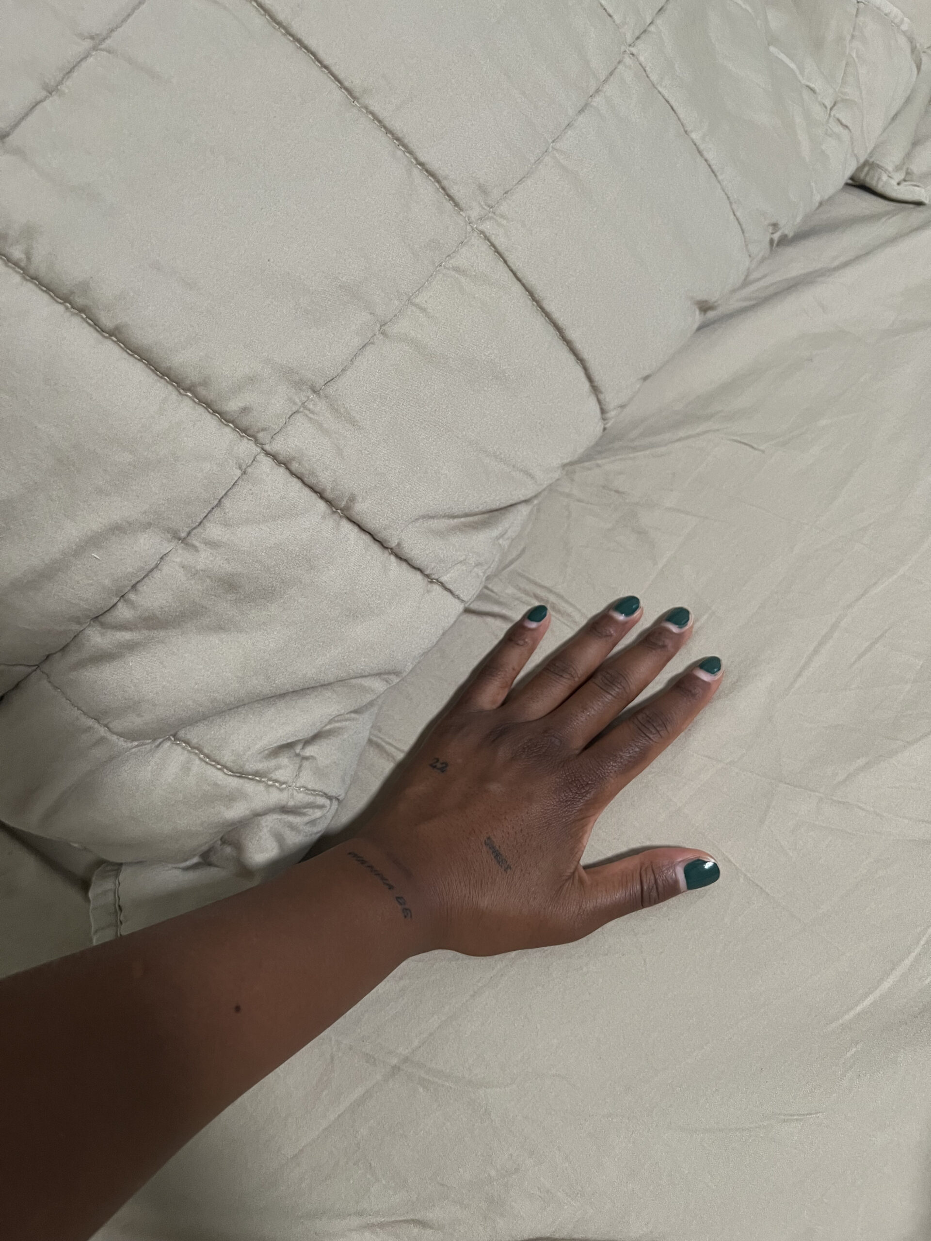 A hand with dark green nail polish rests on a beige quilted blanket.
