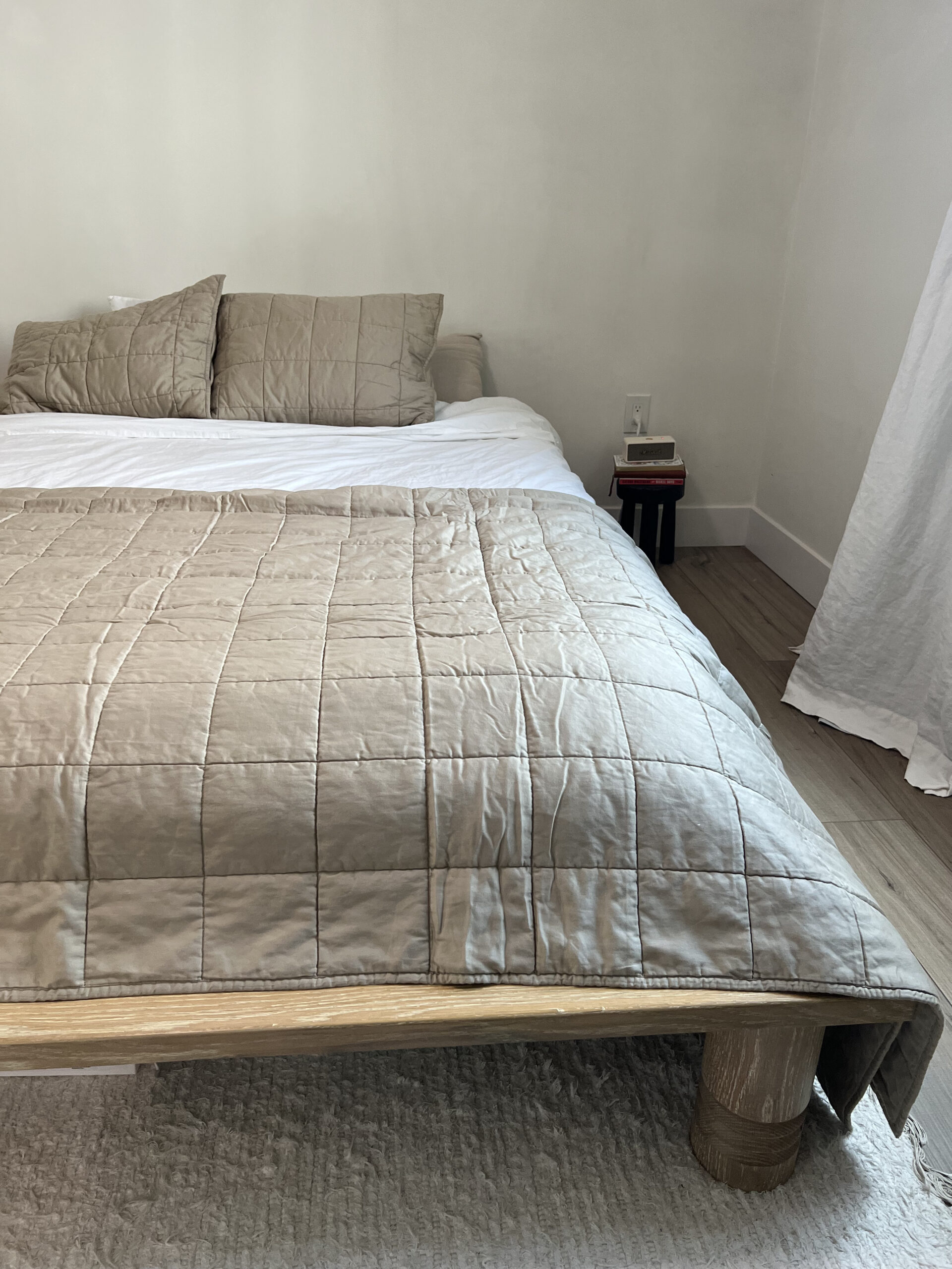 A neatly made bed with a beige quilt, matching pillows, and wooden bed frame. A small nightstand with a book and lamp is visible next to the bed. A white curtain hangs on the right.