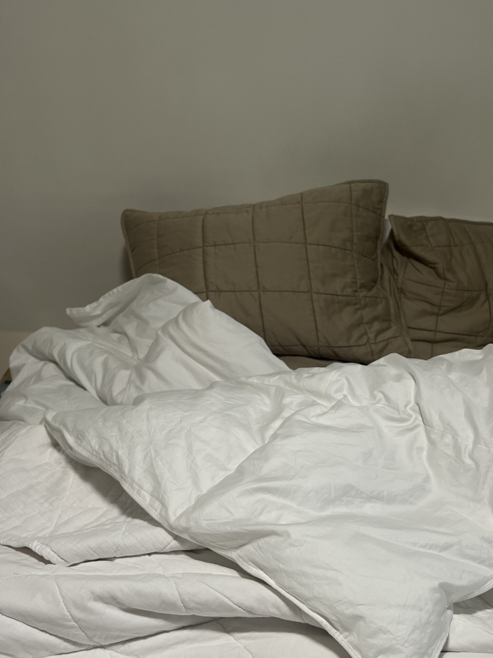 Unmade bed with white sheets and two large brown pillows against a plain wall.