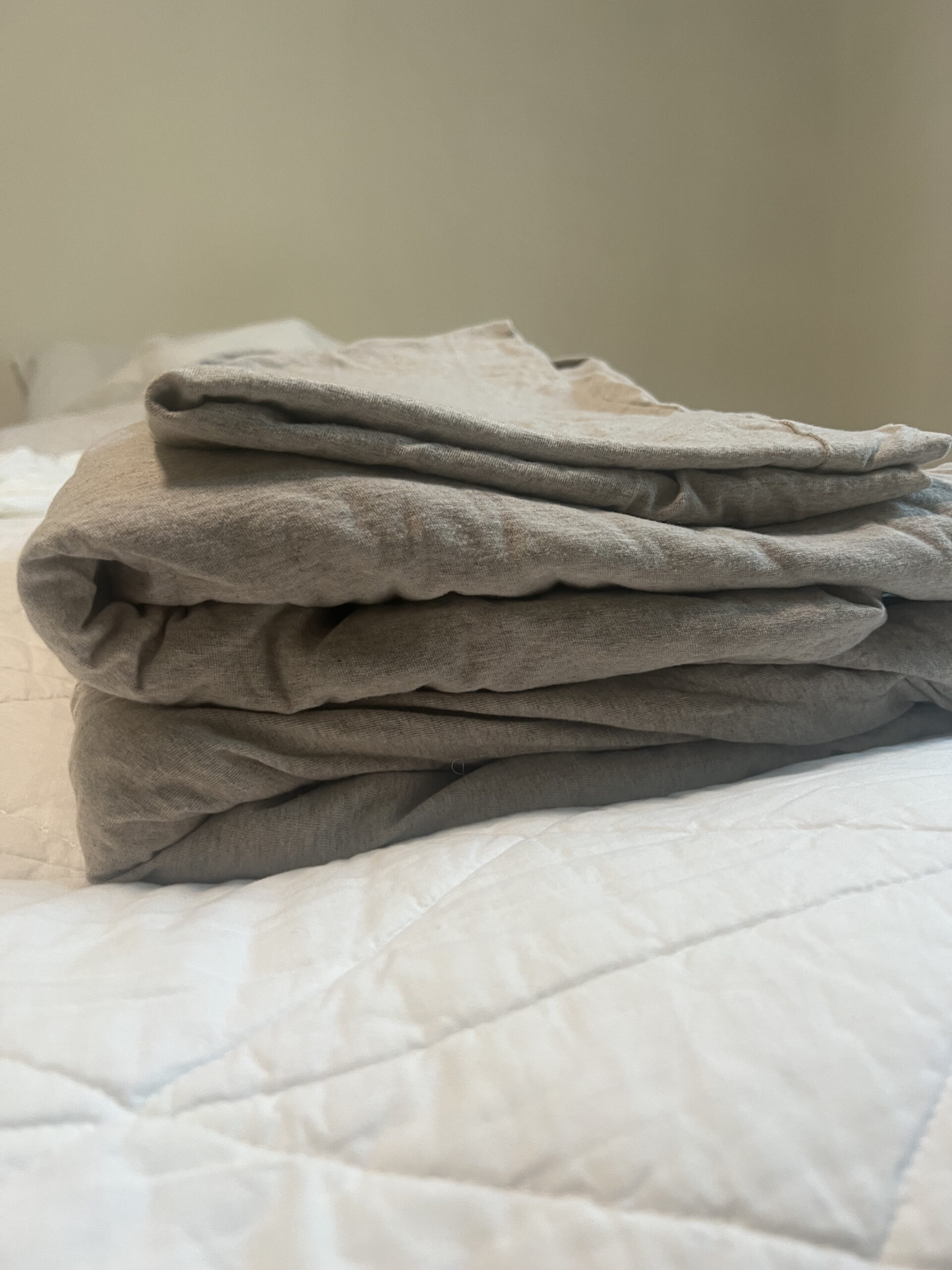 A neatly folded beige blanket sits on a white quilted bedspread in a softly lit room.