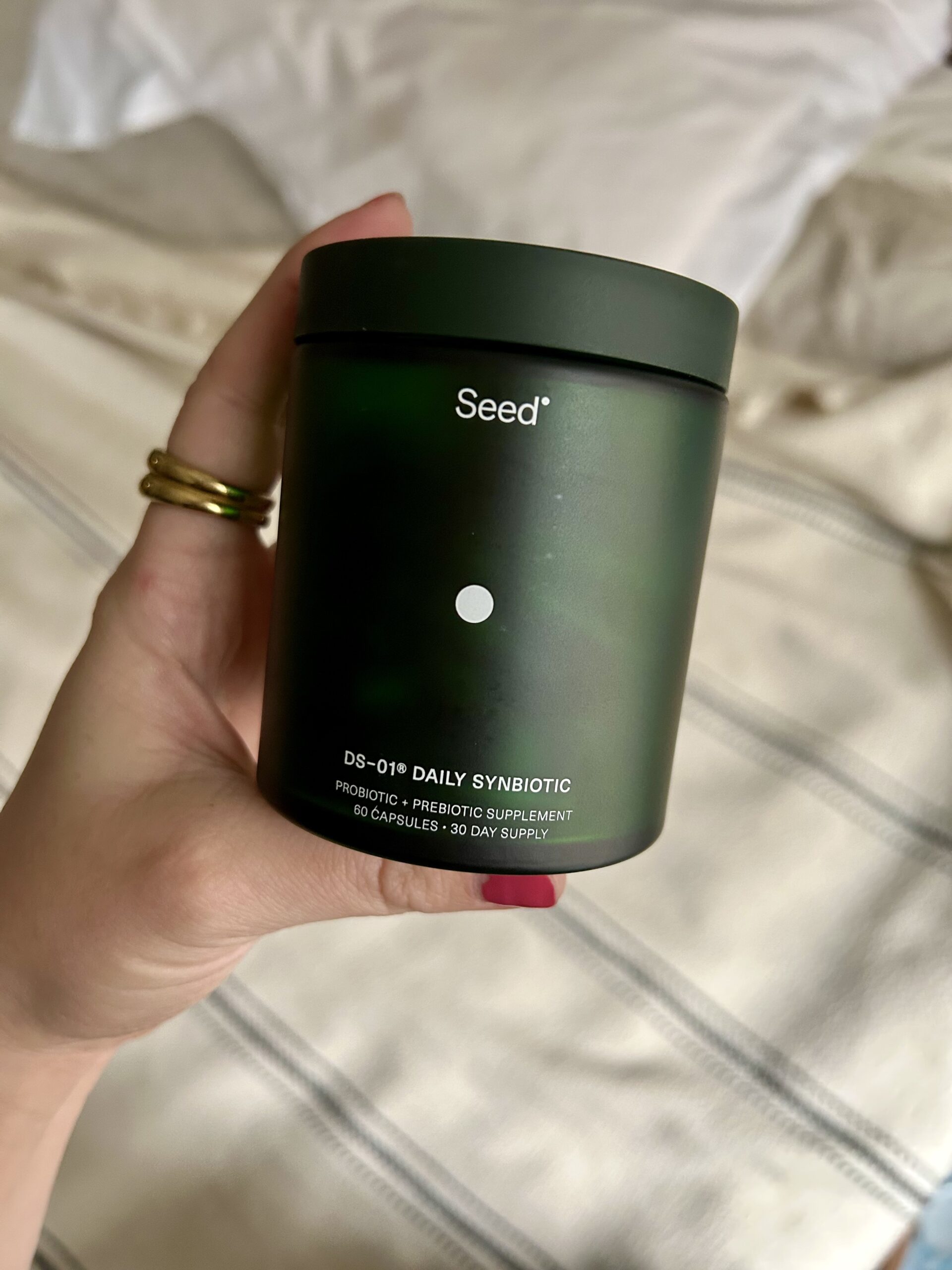 A hand holding a green container labeled "Seed DS-01® Daily Synbiotic" with 60 capsules, described as a probiotic and prebiotic supplement.