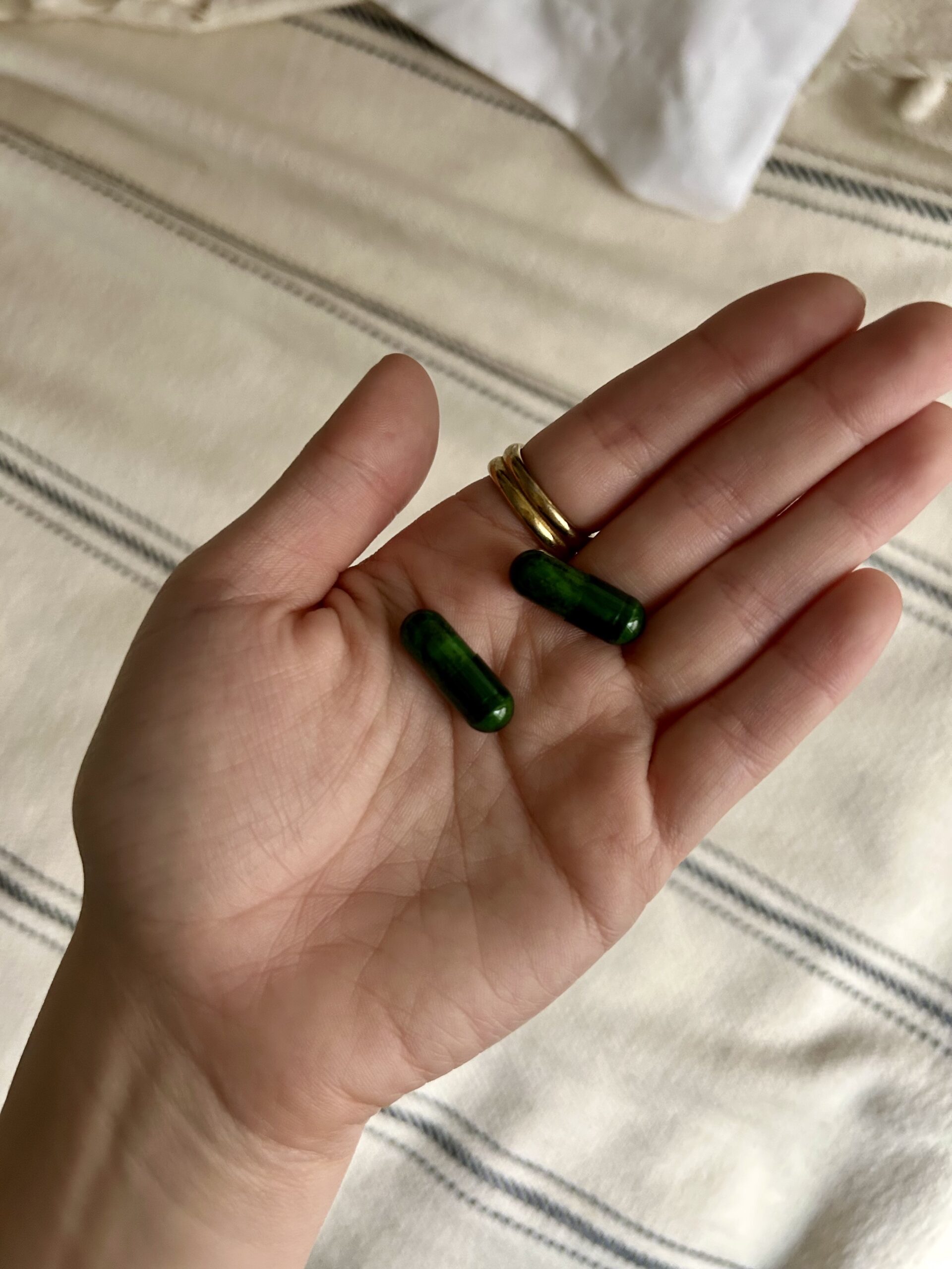 An open hand holding two green capsules, with a beige and striped fabric surface in the background.