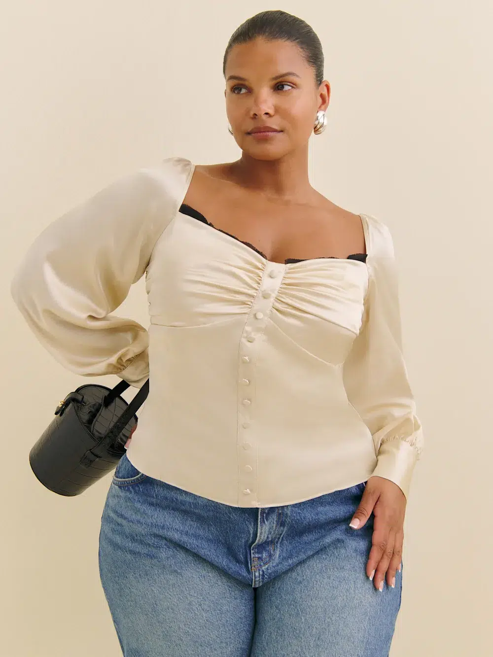 A person in a cream blouse and blue jeans stands against a beige background, holding a small black handbag.