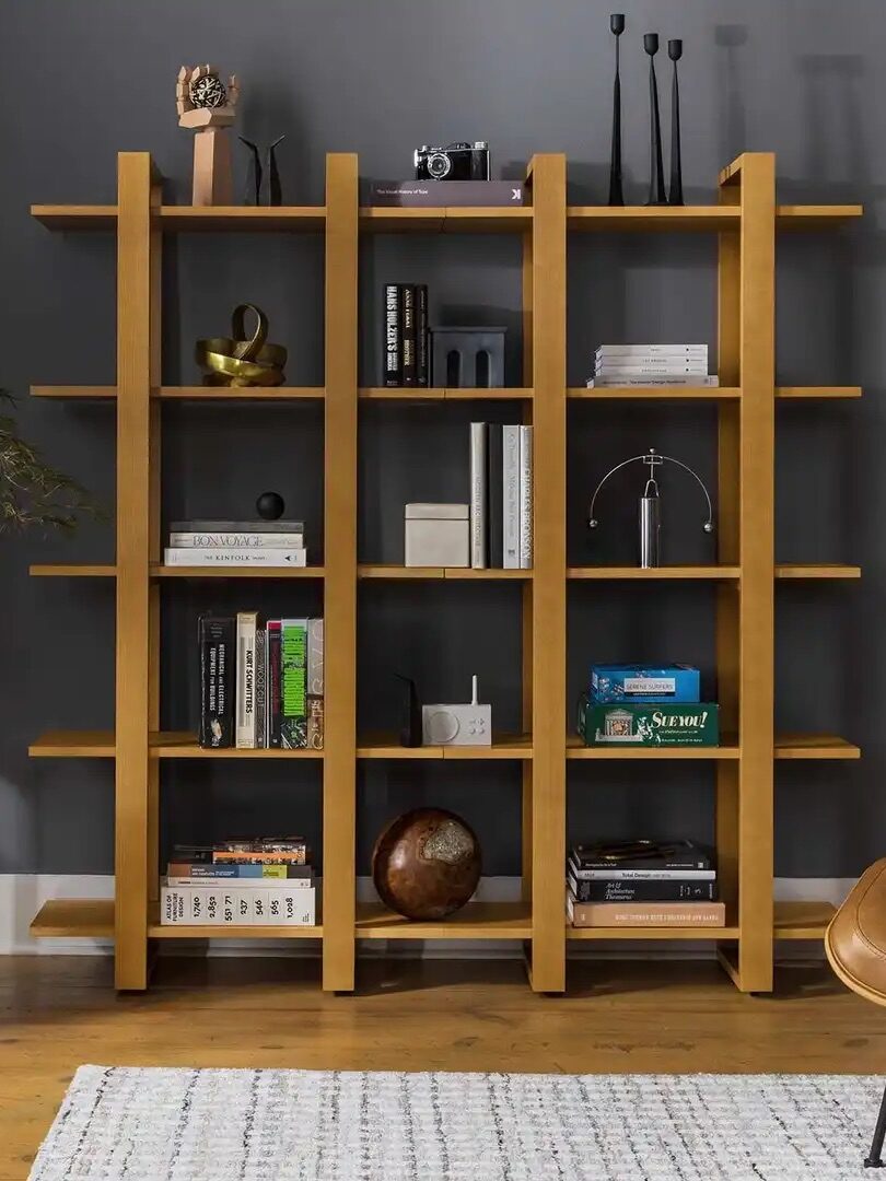 A modern living room with a wooden bookshelf holding books and decor, a small plant on a stool, a leather chair with an ottoman, and a gray wall.