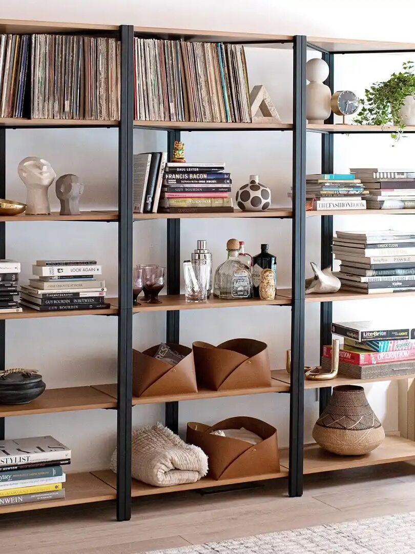 Three-tiered wooden bookshelf with black metal frames, filled with books, plants, records, and decorative items. Nearby is a white speaker with black accents. Large window to the right.