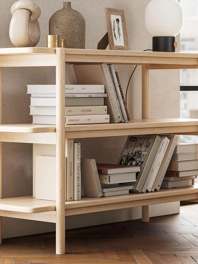 Wooden bookshelf with three shelves, holding various books, vases, and a lamp. Positioned near a window with a green sofa nearby.