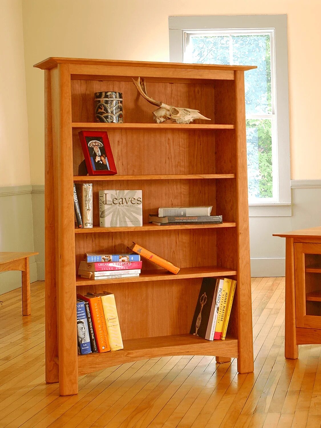 Wooden bookshelf with various books, framed photos, and decorative items beside a wooden cabinet in a room with a window and hardwood floors.