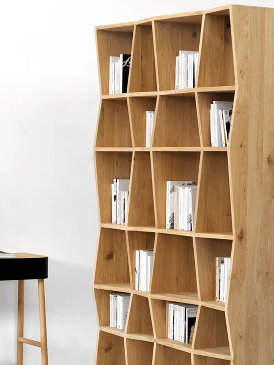 Wooden bookshelf with an irregular design holds books. A small desk and stool are nearby, with an open book on the desk against a plain white wall.