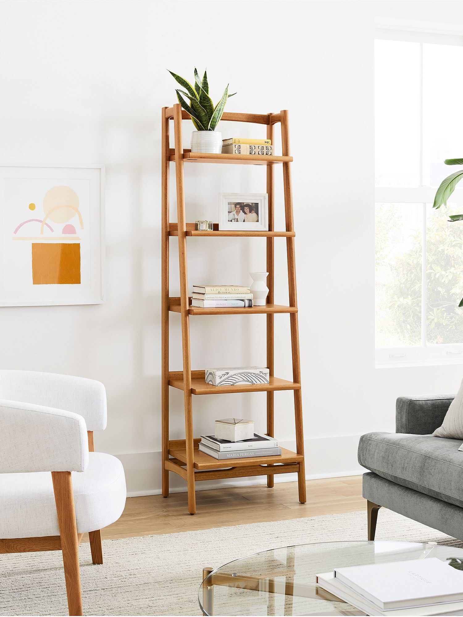 Room interior with a wooden ladder-style shelf holding books and a plant, a white armchair, a gray sofa, framed art on the wall, and a large window allowing natural light.