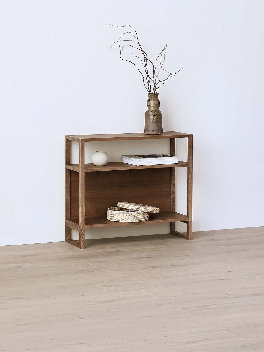 A wooden shelf against a white wall holds a book, a small vase with branches, a round white object, and two woven baskets on the middle shelf.