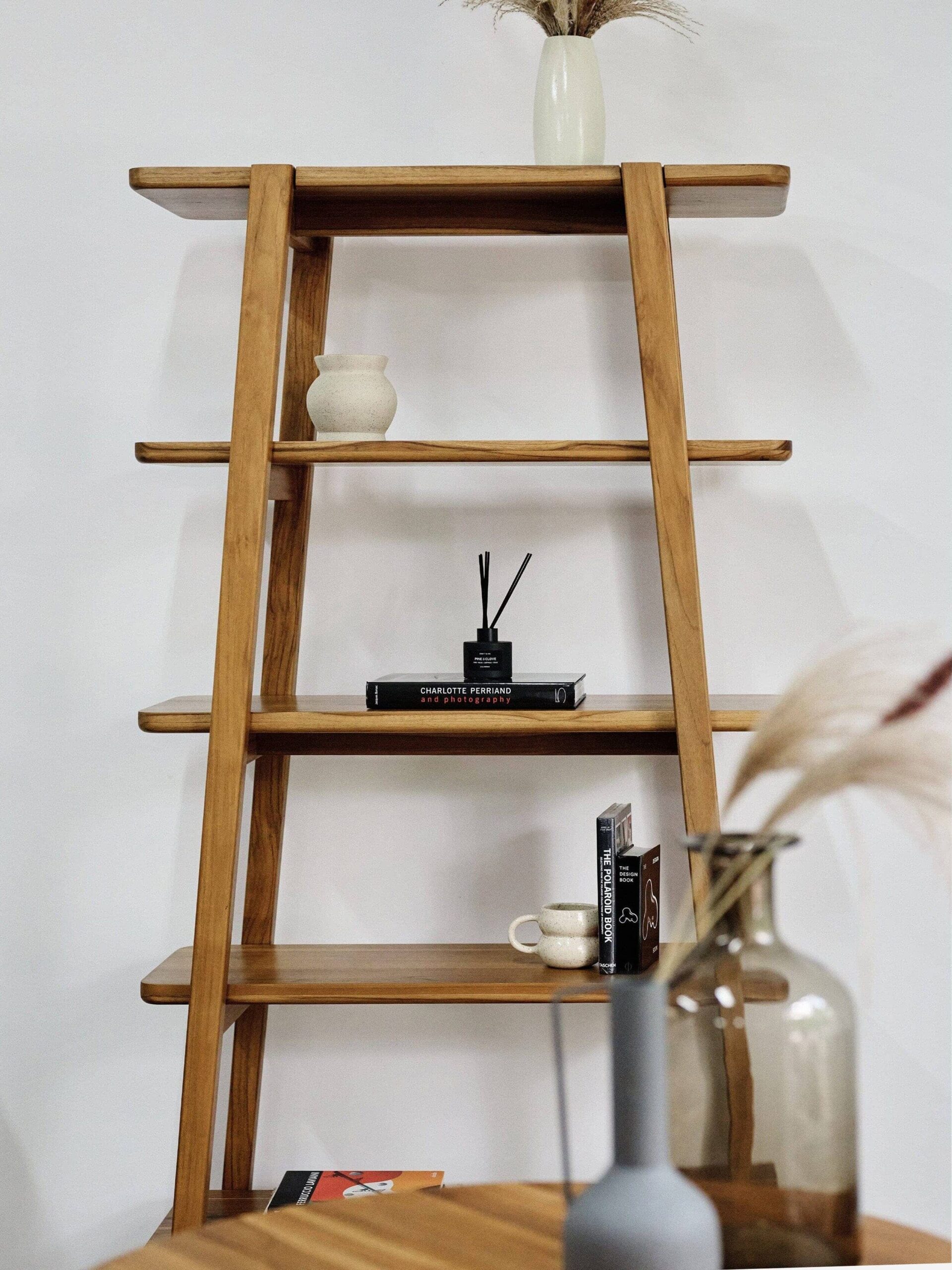 Wooden ladder-style bookshelf with vases, a reed diffuser, books, and a mug placed on its shelves. A small round wooden table with a vase is in the foreground.