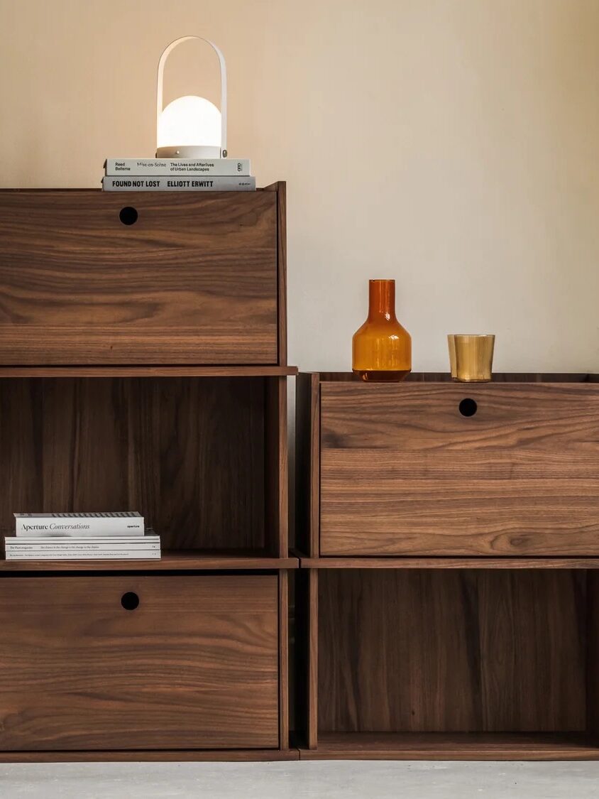 Wooden modular shelving units with a mix of open and closed cubbies. Items include books, vases, a small lamp, and an amber glass bottle on top. A wooden door is on the left.