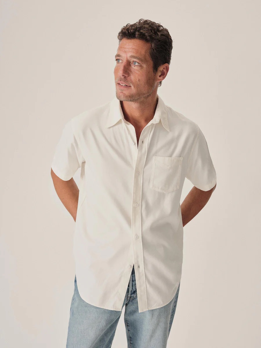 Man wearing a short-sleeve white button-up shirt and light blue jeans stands with hands behind his back, looking to the side against a plain light background.