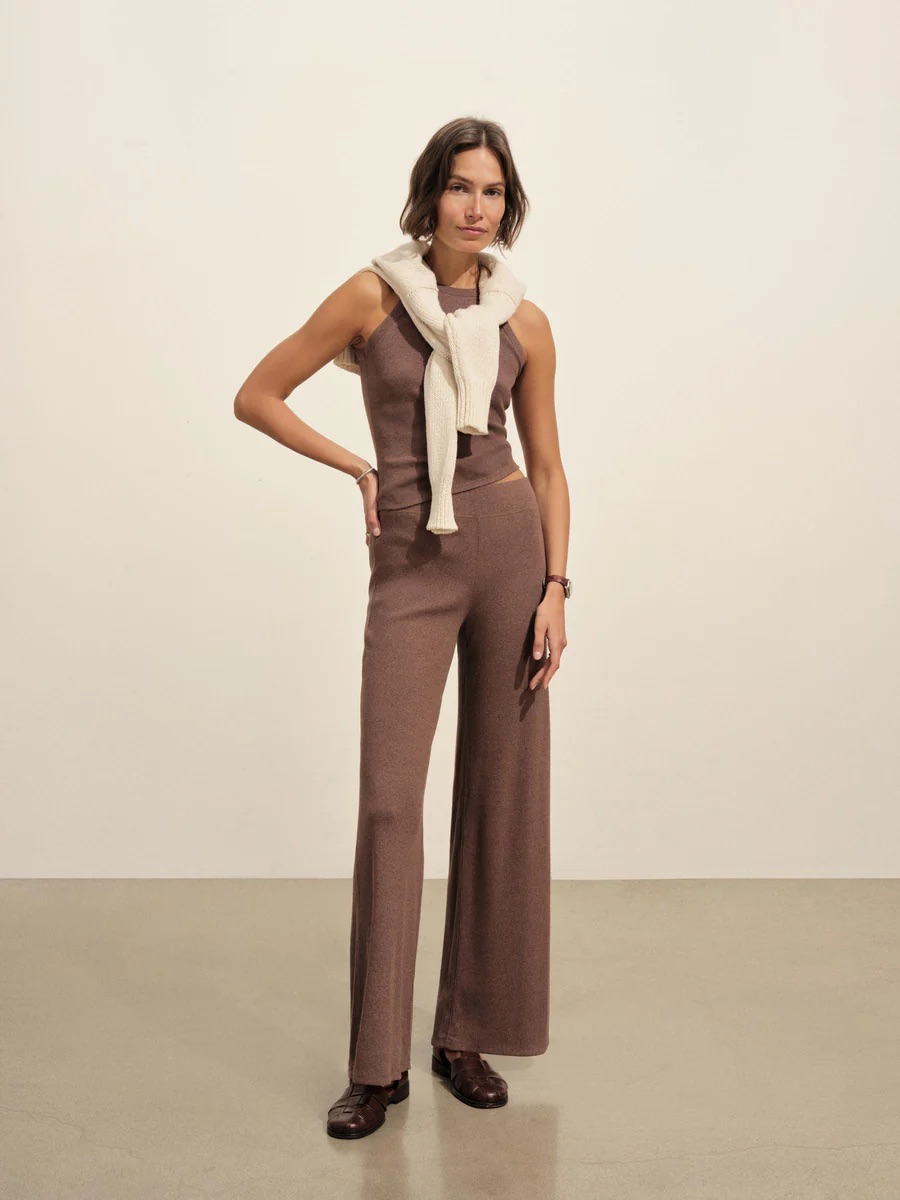 A woman stands indoors wearing a sleeveless brown knit jumpsuit, a cream sweater draped over her shoulders, and brown shoes.