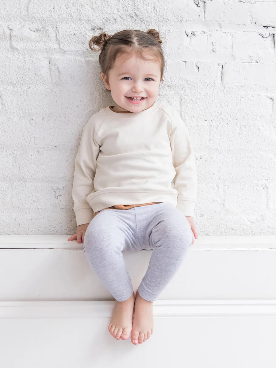 A young child with light brown hair in two buns, wearing a cream sweatshirt and gray leggings, sits barefoot against a white brick wall, smiling at the camera.