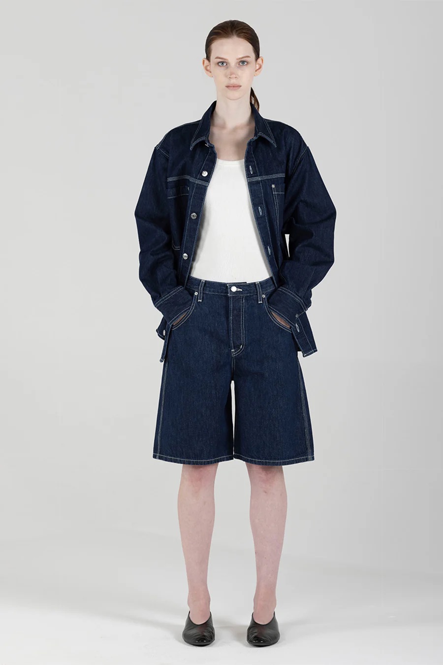 A person stands facing forward wearing a dark denim jacket, matching long denim shorts, a white top, and black shoes against a plain background.