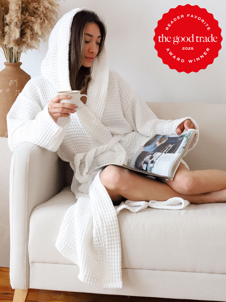 A woman in a white robe sits on a couch reading a magazine and holding a mug. A red “The Good Trade 2025 Award Winner” badge is in the upper right corner.