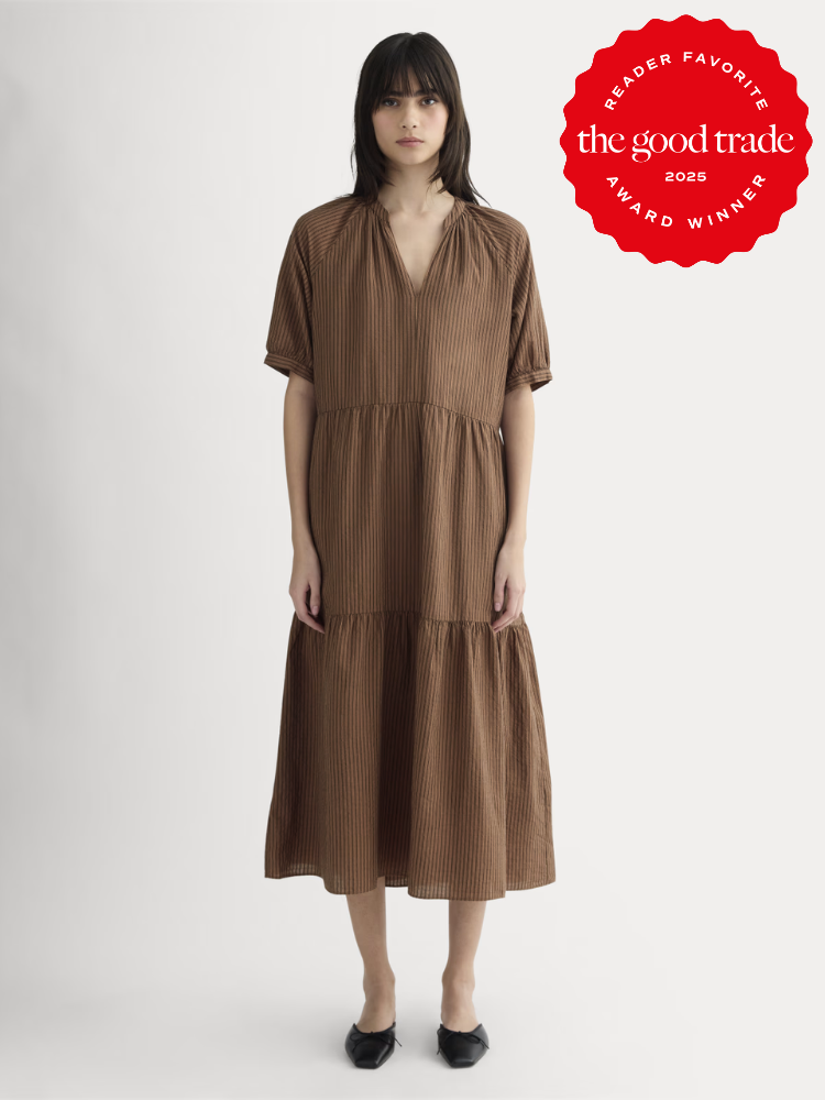 A woman stands facing forward wearing a loose-fitting, brown striped midi dress with short sleeves and black flats. A "The Good Trade 2025 Award Winner" badge is in the top right corner.