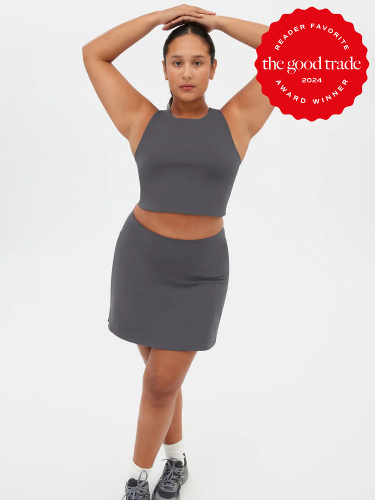 A person poses in a dark gray athletic crop top and skirt set with sneakers. A red badge in the corner reads, “the good trade award winner 2024 reader favorite.”.