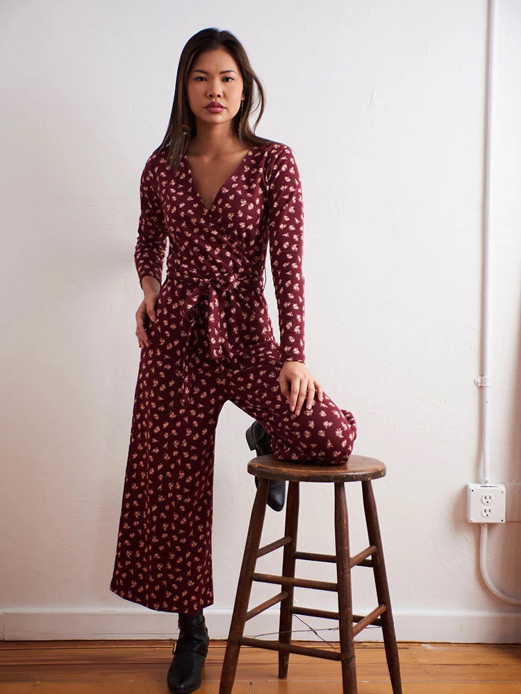 Woman wearing a maroon patterned jumpsuit stands indoors with one leg resting on a wooden stool against a plain white wall.