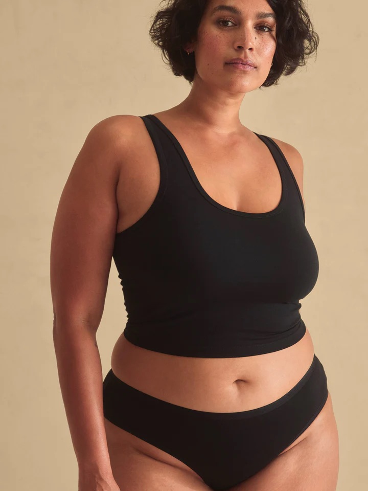 A woman with short curly hair wears a black tank top and black underwear, standing against a plain beige background.