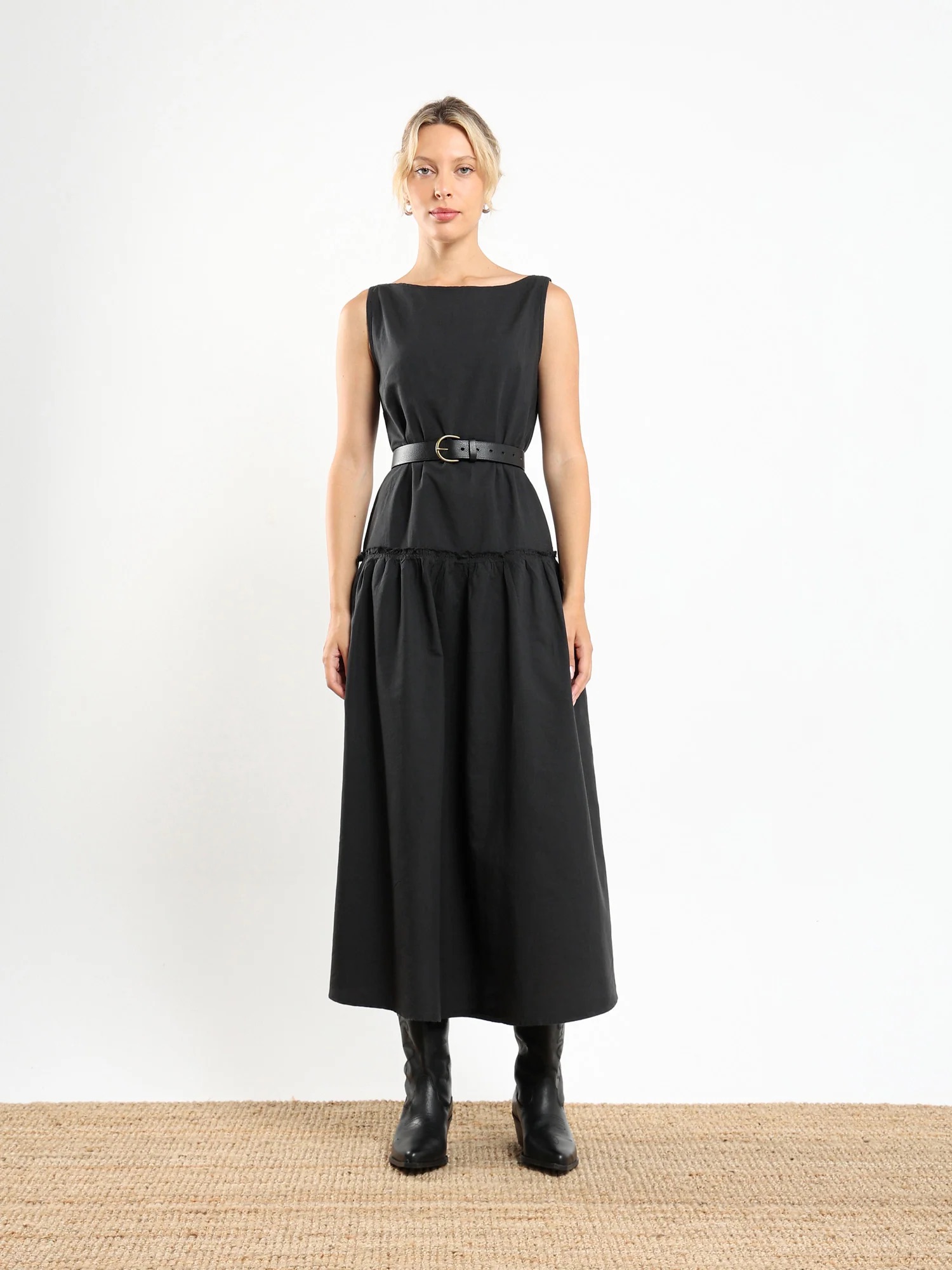 A woman stands facing forward wearing a sleeveless black dress with a belt and black boots, against a plain white background on a woven floor mat.