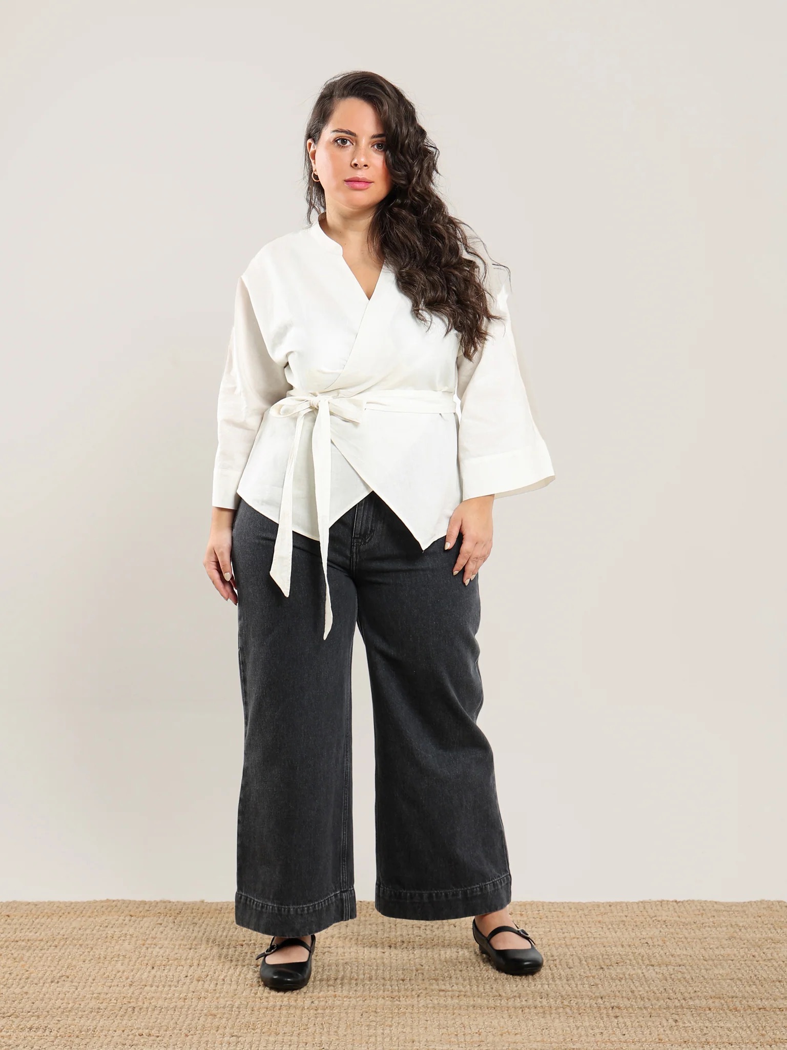 Woman standing on a woven rug wearing a white wrap top with a belt, wide-leg black jeans, and black shoes, against a plain light background.