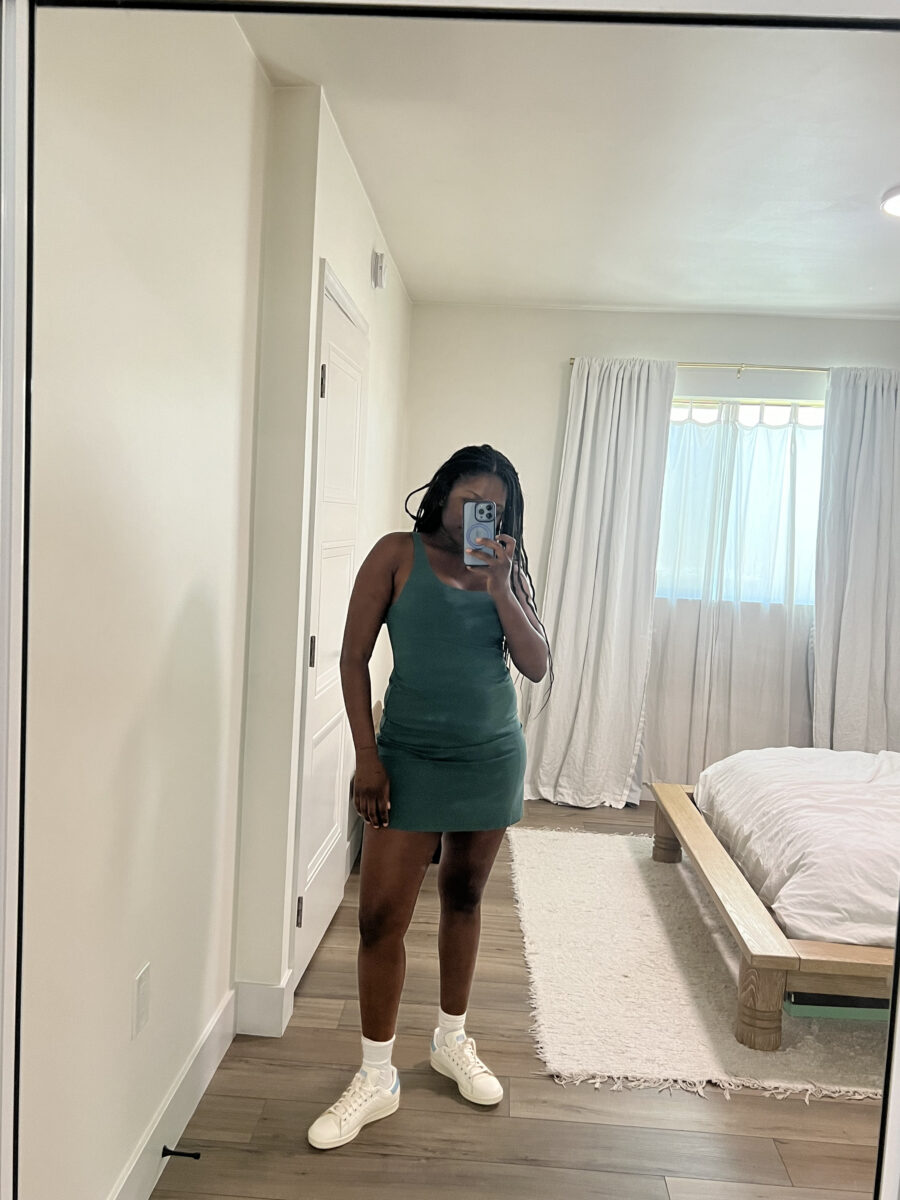 A person stands in front of a mirror taking a selfie, wearing a green sleeveless dress and white sneakers, in a bedroom with light-colored walls and furniture.